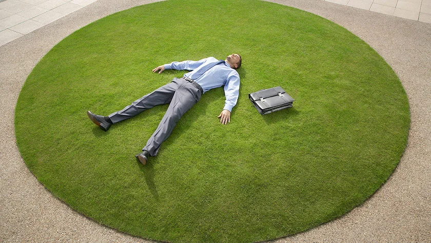 A man lying on his back on a patch of grass. His briefcase is next to him.